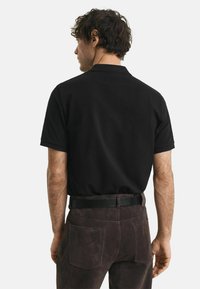Man met krullend haar van zich af gericht, gekleed in een zwart polo T-shirt met korte mouwen, ingetrokken in een donkerbruine pantalon met een zwarte riem.
