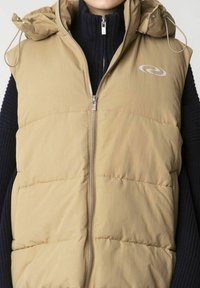 Tan puffer-väst med dragkedja framtill, huva och kviltad design. Har en logotyp och är lager-på-lager över en mörk, strukturerad tröja.