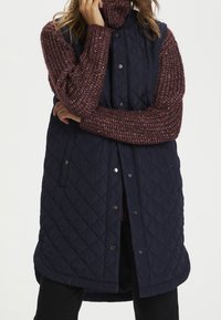 Gilet imbottito blu navy con bottoni a pressione e tasche laterali, indossato sopra un maglione a maglia borgogna con pattern testurizzato. Taglio lungo e relax.