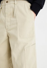 Beige cargo pants made of lightweight cotton fabric, featuring side pockets and a relaxed fit. Visible stitching and button closure at waistband.