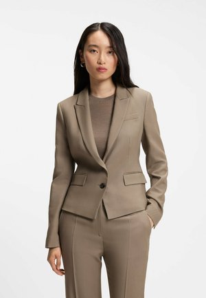 Kvinde iført en tætsiddende taupe blazer og matchende bukser med en brun top med rund hals, står med den ene hånd i lommen mod en hvid baggrund.