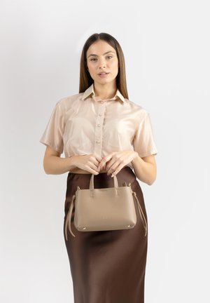 Donna con capelli castani lunghi che indossa una camicia beige in raso a maniche corte e una gonna marrone scuro, che tiene una borsa in pelle color taupe davanti a uno sfondo bianco.