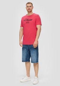 Man in een rode s.Oliver t-shirt, blauwe denim knielange shorts en witte sneakers, staand tegen een effen lichte achtergrond.