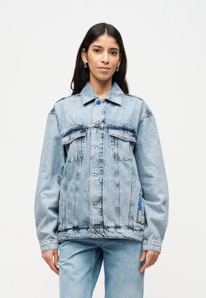 Woman wearing light blue oversized denim jacket and matching jeans, standing against a plain white background.