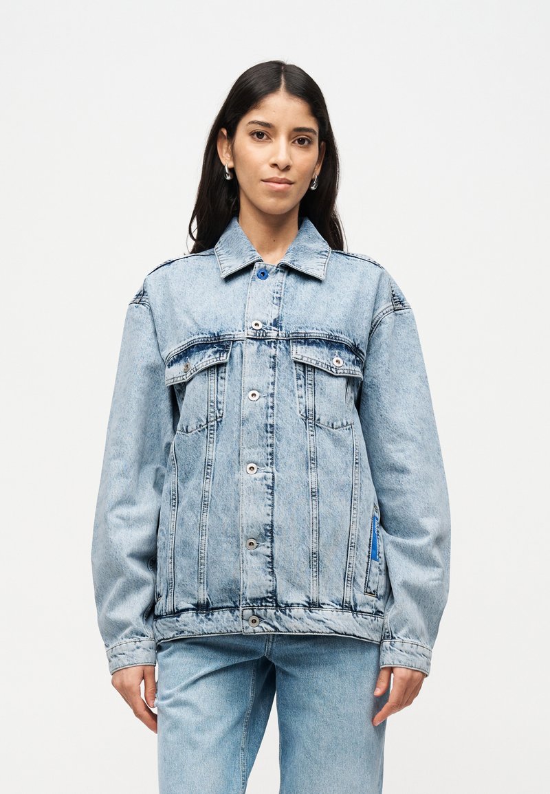 Γυναίκα που φοράει ανοιχτό μπλε υπερμεγέθη τζιν μπουφάν και τζιν παντελόνι που ταιριάζουν, στέκεται μπροστά σε απλό λευκό φόντο.