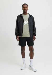 Chaleco cortavientos negro con cremallera, camiseta verde oliva con logotipo blanco, pantalones cortos negros, zapatillas blancas y calcetines blancos con logotipo.