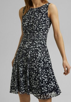 Robe noire sans manches avec motif floral blanc, taille ajustée et jupe évasée portée par une femme sur un fond gris uni.