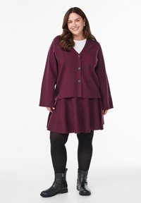 Uśmiechnięta kobieta w bordowej kardiganie zapinanym na guziki oraz dopasowanej spódnicy, w czarnych rajstopach i czarnych botasach za kostkę, stojąca na tle białego tła.