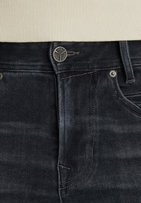 Tamne traperice tamnoplave koje sadrže klasičan dizajn, srebrne detalje i suptilnu istrošenost na pojasu i džepovima.