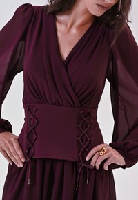 Bordeaux jurk met een wikkeldesign, doorzichtige lange mouwen en veterdetails op de taille. De stof is soepel met een subtiele glans.