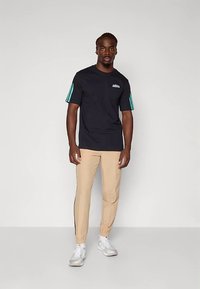 Černé tričko s tyrkysovými pruhy na rukávech, s logem na hrudi, kombinované s béžovými tepláky se bočními pruhy, nošené s bílými sportovními botami.