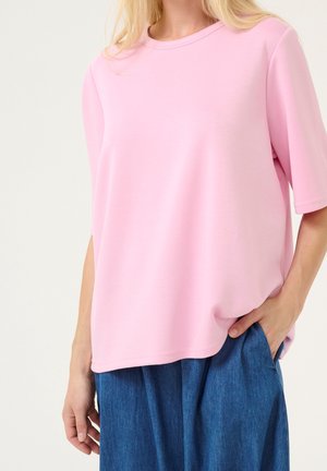 Personne portant une chemise ample à manches courtes rose clair et un pantalon bleu à jambes larges, une main dans la poche, sur un fond uni.