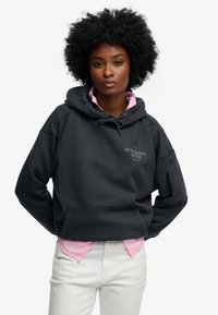 Mørkegrå sweatshirt med hætte, der har en frontlomme og en hvid logo. Bruges over en lyserød skjorte og kombineret med hvide bukser.