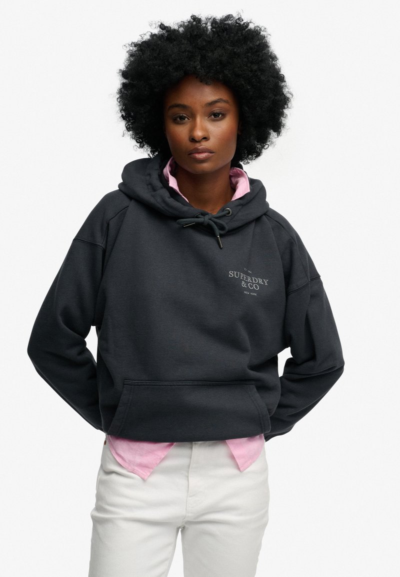 Mørkegrå sweatshirt med hætte, der har en frontlomme og en hvid logo. Bruges over en lyserød skjorte og kombineret med hvide bukser.