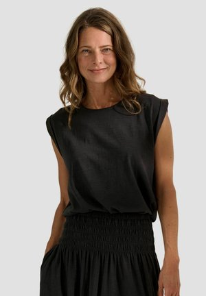 Donna con capelli castani ondulati che indossa un top nero senza maniche e una gonna nera a vita alta, sorridente, con una mano in tasca.