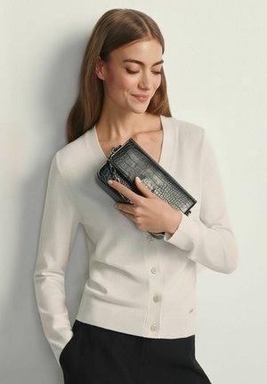 Młoda kobieta w białym swetrze na guziki, trzymająca czarną kopertówkę z tłoczeniem imitującym skórę krokodyla, delikatnie się uśmiecha z zamkniętymi oczami.