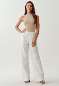 Beež ilma varrukat crop-top kõrge kaelusega, kombineeritud valgete laia jala pükstega, millel on puhas disain ja minimaalsed aktsendid.