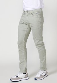 Jeans de denim verde claro con un ajuste slim, que cuentan con una cierre frontal de botón y cremallera, bolsillos laterales y bolsillos traseros. Combinados con zapatillas blancas y grises.