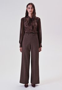 Bruine doorschijnende bloes met versieringen en strik, gecombineerd met wijde geruite bruine broek en bordeauxkleurige schoenen. Gladde textuur en op maat gemaakte pasvorm.