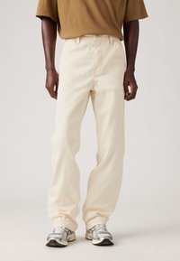 Pantalones de mezclilla beige con un corte relajado, que presentan una cintura alta, bolsillos frontales y un cierre de botón metálico. Combinados con zapatillas grises.
