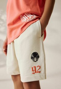 Jasnopomarańczowa koszulka zestawiona z kremowymi szortami zdobionymi czarną grafiką goryla oraz pomarańczowym nadrukiem "92" na nodze. Casualowy materiał i luźny krój.