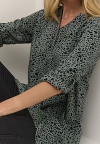 Blusa verde con manchas de leopardo negras, fabricada en un tejido ligero, con un corte ligeramente holgado, escote en V y mangas enrolladas.