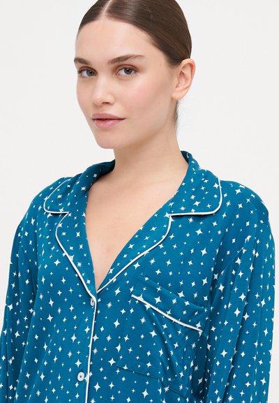 Camisa de pijama azul-petróleo com padrão de estrelas brancas, colarinho com recorte e fecho de botão. Apresenta um bolso no peito com borda branca. Tecido suave e leve.