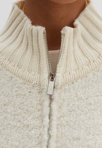 Vaalea beige vetoketjullinen takki, jossa on ribbineulottu korkea kaulus, teksturoitu kangas ja hopeinen vetoketju. Takissa on pehmeä, mukava ilme ja neulottu viimeistely.