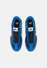 Zapatillas azules y negras con materiales de ante y cuero, que presentan detalles amarillos, cordones negros y una etiqueta de logo blanca en la lengua.