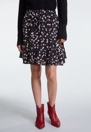 Kvinde iført en sort blomsterprint nederdel med flæsekant, sort langærmet trøje og røde cowboystøvler, stående foran en grå baggrund.