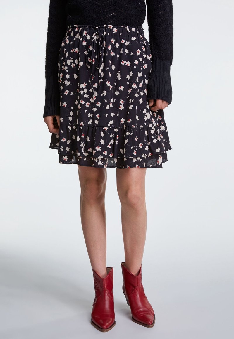Femme portant une jupe noire à motif floral avec un ourlet à volants, un pull noir à manches longues et des bottines rouges de style cowboy, debout devant un fond gris.