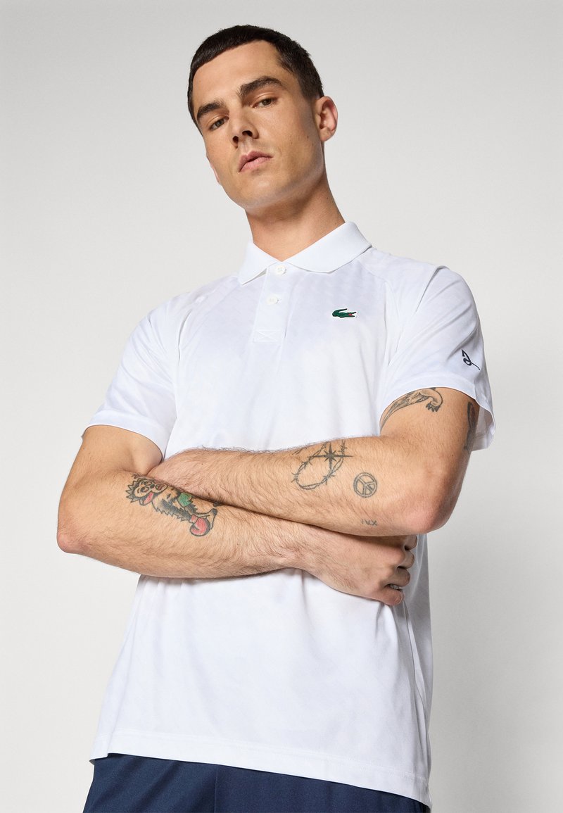 Polo shirt blanc en tissu respirant, avec un logo vert sur la poitrine, des manches courtes et un design de col classique.