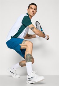 Conjunto de ténis masculino: camisa de mangas curtas branca e teal, calções azuis, sapatilhas brancas e meias brancas. Apresenta tatuagens visíveis nas pernas.