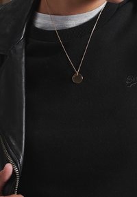 Collar de colgante circular en tono dorado sobre una cadena fina, llevado sobre un suéter negro, complementado con una chaqueta de cuero negra.