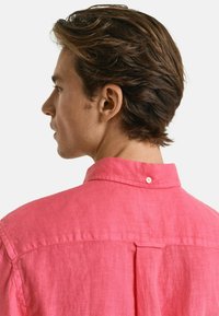 Rosa Leinenhemd mit klassischem Kragen, Rückenfalten und einem einzelnen Knopfverschluss am Hals, das einen glatten, strukturierten Stoff zeigt.