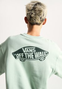 Sudadera en verde menta con un gran logo negro de "Vans 'Off the Wall'" en la parte posterior, con una textura suave y redondeada en el cuello.