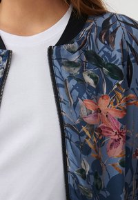 Květinová bomber bunda z modré látky, zdobená barevnými květinami a listy. Má černý límec a zip, vrstvená přes bílý top.