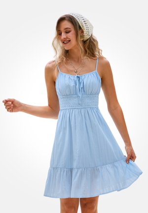 Smiling woman wearing a light blue sleeveless dress and white knitted headscarf, holding dress hem with one hand against plain background.