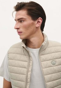Beige polstret vest med høj krave, med vandret syning, lynlås foran og et rundt logo-patch på venstre bryst. Bæres over en grå T-shirt.