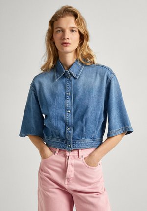 Giacca in denim blu tagliata, con maniche corte, chiusura a bottoni e una texture leggermente rovinata, abbinata a jeans rosa chiaro a vita alta.