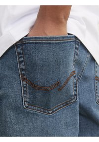 Mano insertada en el bolsillo trasero de unos jeans de mezclilla azules con costuras naranjas, vistiendo una camisa blanca.