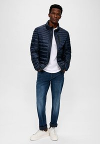 Marineblauwe geïsoleerde jas met een rits aan de voorkant, gecombineerd met een wit T-shirt en donkerblauwe jeans. Witte sneakers maken de outfit compleet.