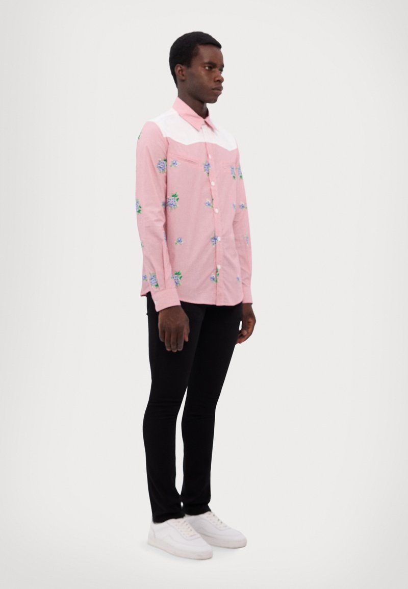 Chemise boutonnée rose avec un motif floral bleu et vert, col contrastant blanc, manches longues, associée à un pantalon slim noir et des baskets blanches.