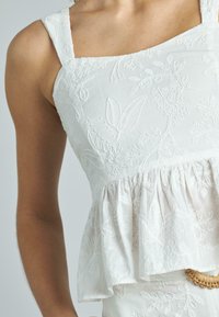 Top bianco a peplo con motivi floreali ricamati, scollatura quadrata e spalline larghe. Tessuto morbido, leggero e dettagli in vita arricciati.