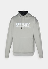 Šviesiai pilka sportinė striukė su juodu vidiniu apykakle, su baltu "OAKLEY Thermonuclear Protection" logotipu ir priekyje esančia kišene. Minkšta medžiaga, laisvas siluetas.