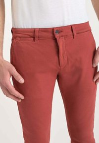 Man som bär slim-fit byxor i rostfärg med knapp och dragkedja, tillsammans med en vit skjorta som är croppad vid midjan och låren.