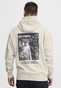 Sweat à capuche beige avec un graphique au dos présentant une image texturée en noir et blanc d'un quai de gare et du texte. Matière en coton, coupe décontractée.