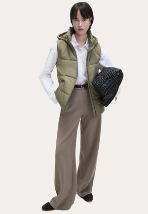 Oliwkowa kamizelka puchowa z kapturem, noszona na białej koszuli; w zestawieniu z beżowymi spodniami o szerokich nogawkach i czarną torebką o fakturze.