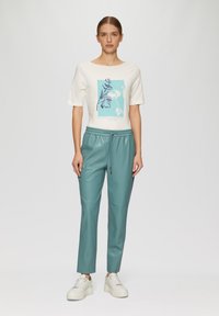 Weißes kurzärmeliges Hemd mit blauem Grafikdesign, kombiniert mit tealfarbenen Kunstlederhosen. Das Model trägt weiße Sneaker. Lässiges Outfit.