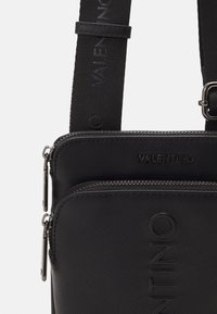 Černá texturovaná crossbody taška s vyraženým logem Valentino, dvojitými zipovými přihrádkami a nastavitelnou přezkou na ramenním popruhu s logem.